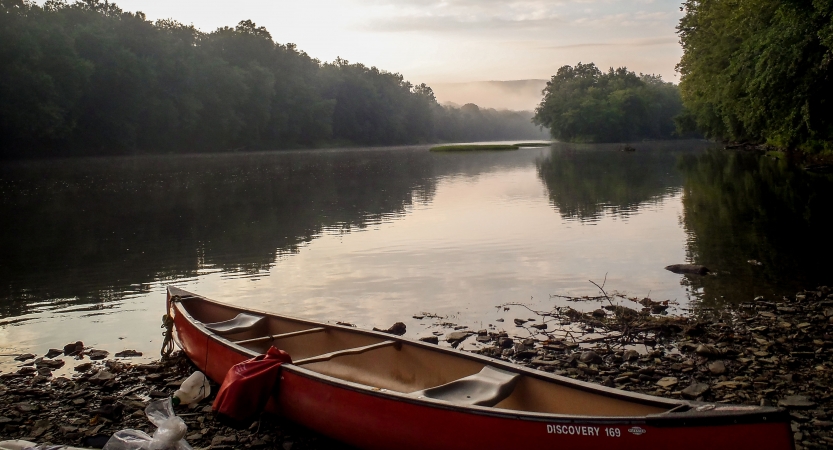 A canoe rests on a rocky shore beside a river lined with green trees. 